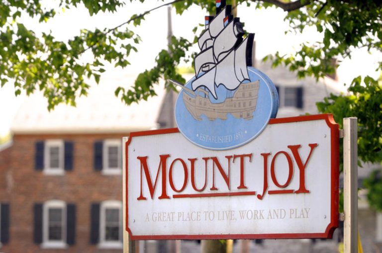 Sign for Mount Joy, featuring a ship illustration and the words “Established 1851” above the town name in large red letters. Below, the sign reads “A great place to live, work and play.” A brick building and green tree leaves are visible in the background.